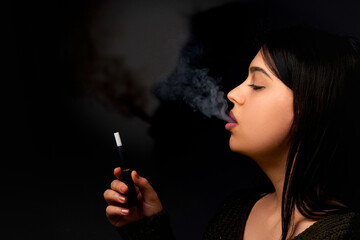 Fototapeta premium A young woman smokes a vape and lets out smoke on a dark background, Vaping concept with place for text.