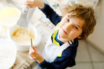 Adorable funny blond little kid boy baking chocolate cake and tasting dough in domestic kitchen, indoors. Happy child having fun with working with mixer, flour, eggs dough at home. Little helper