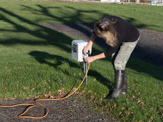  A female motorhomer plugs the orange electric hook up lead laying on floor into a campsite supply point using the blue connector.She wears black rubber boots and camouflage trousers on sunny day with - obrazy, fototapety, plakaty
