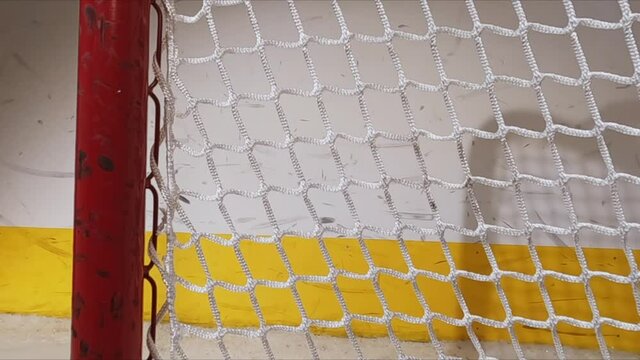 A close-up view towards the inside of the goal net as an Ice Hockey puck hits the back of it as shavings fly by, then bounces back at the camera. Scoring a goal in ice hockey.
