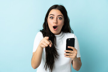 Fototapeta premium Young woman using mobile phone isolated on blue background surprised and pointing front