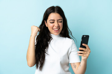 Young woman using mobile phone isolated on blue background celebrating a victory