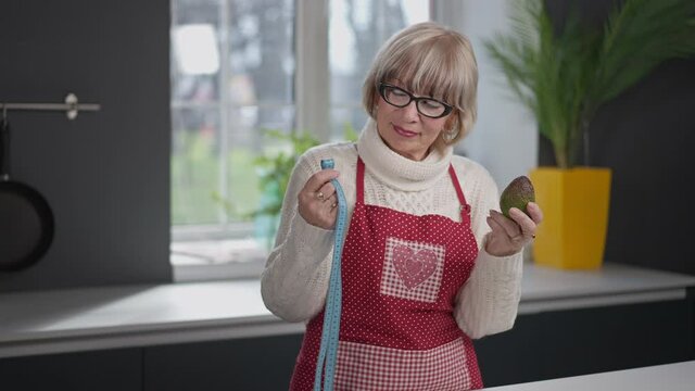 Happy beautiful senior woman with avocado and measuring tape smiling talking looking at camera. Portrait of slim gorgeous Caucasian retiree in eyeglasses posing indoors at home. Healthy eating concept
