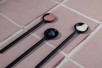 Three black spoons with cream smear or black and white mud mask smudge texture on pink tile bathroom floor bathroom with copy space. Natural beauty skin care cosmetic product.  Flat lay. Facial spa.