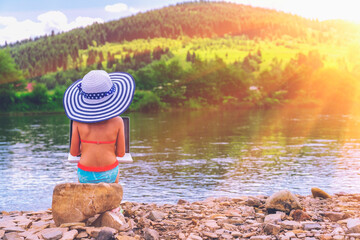 Young girl dressed in a straw beach sun hat working or studying remotely with a laptop outdoors on the river in front of the mountains
