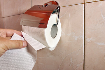 The man unrolls some toilet paper from the holder and tears it off. The concept of personal hygiene.