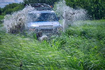 Obraz na płótnie Canvas Off-road car crashed into a puddle and raised mud splashes. A four-wheel drive sports car is racing among the green grass. Off-road 4x4 SUV in blue color. The best off-road vehicles