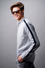 Portrait of smiling handsome guy standing in sunglasses holds arms in pockets, isolated on a gray background