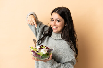 Young caucasian woman isolated on beige background with happy expression while pointing a bowl of salad