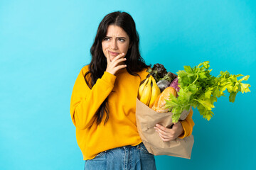 Young woman holding a grocery shopping bag isolated on blue background having doubts while looking...