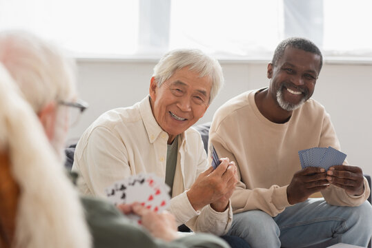 Elderly multiethnic men playing cards with blurred friend at home.