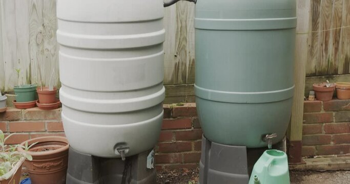 A down-pipe going into a large green water barrel which is joined with another one for more water storage. Camera tilts down.