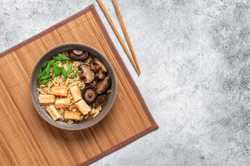 Asian vegan ramen soup with noodles, tofu cheese and shiitake mushrooms in a bowl on a bamboo mat. Gray concrete grunge background. Top view.