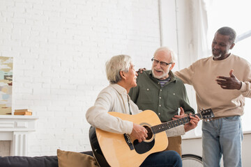 Positive senior friends looking at asian man playing acoustic guitar at home.