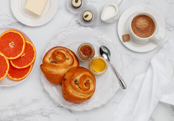 Two buns with raisins, jam and honey in a white plate on a marble background for breakfast. Coffee, milk, butter,  chocolates and oranges on the table. Selective focus, top view