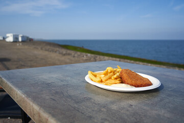 White plate with fast food on the beach. Schnitzel and fries with sauce on the table. Sea water and...
