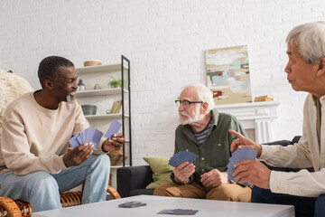 Smiling african american man holding playing cards near multiethnic friends in living room.