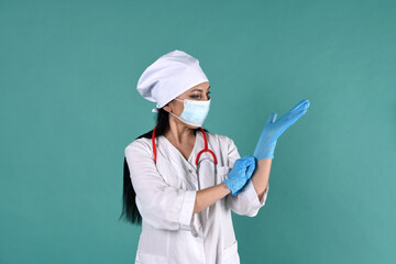 A female doctor in a white coat and a white headdress does puts on blue medical gloves on a green background with a stethoscope