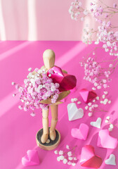 a wooden man holding a heart and a bouquet on a pink background surrounded by flowers and hearts

