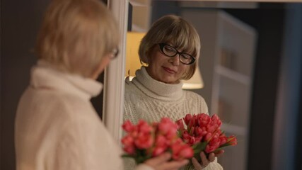 Reflection in mirror of senior woman with bouquet of flowers standing indoors. Beautiful female Caucasian retiree admiring red tulips in living room at home. Birthday or Women's Day celebration