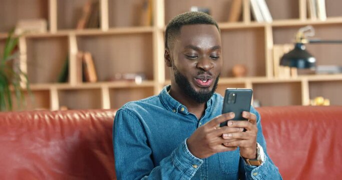 Young African American man in denim shirt is resting on red sofa in modern office during break and spends time on social media using smartphone.