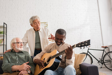 Cheerful african american man playing acoustic guitar near multiethnic friends at home.