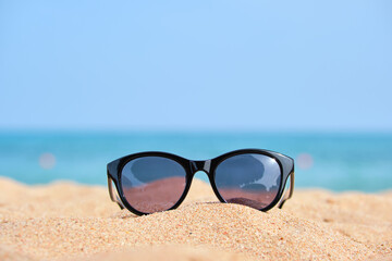 Fototapeta na wymiar Closeup of black protective sunglasses on sandy beach at tropical seaside on warm sunny day. Summer vacation concept