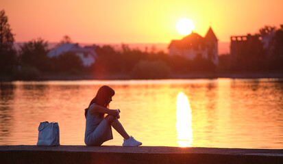 Lonely young woman sitting alone on lake shore enjoying warm evening. Wellbeing and relaxing in nature concept