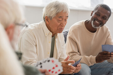 Upset asian man playing cards with multiethnic friends at home.