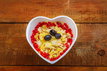 Puff salad with chicken breast and pineapple, garnished with pomegranate in a heart salad bowl on a wooden table.