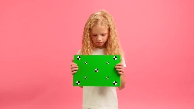 Happy Blonde girl holding Banner with Green Screen Tracks Points for Copy Space. Empty Green Screen Board. Smiles and Looks at Camera With Space for Text or Advertising on Pink Background.