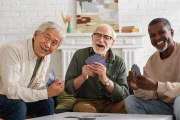 Cheerful interracial friends holding playing cards and looking at camera at home.