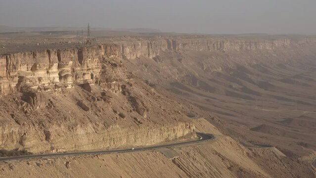 Car drives past dramatic ridge of the Makhtesh Ramon, a crater formed by erosion in the Negev desert. Natural landscape and transportation in Israel.
