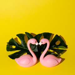 Two birds, pink flamingos kissing and making a silhouette of a heart in front of two palm leaves on a bright yellow background
