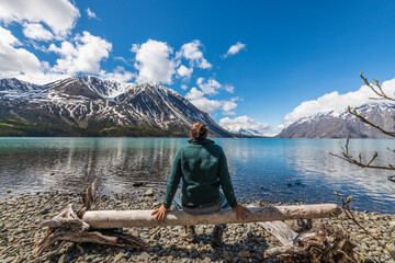 Tourist, hiker sitting beside stunning lake in northern Canada during summer time with snow capped mountains in background, pristine lake and rocky lakeshore. 