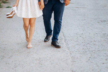 A couple of lovers, a man and a woman, the bride and groom are walking along the road holding hands. The woman took off her high-heeled shoes, goes barefoot, her legs are tired. No face.