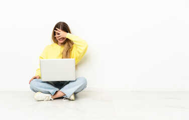 Young woman with a laptop sitting on the floor covering eyes by hands and smiling