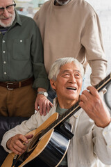 Elderly asian man playing acoustic guitar near interracial friends at home.