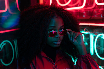 Millennial enigmatic pretty girl with unusual dyed hairstyle near glowing neon wall at night. Blue hair, golden sequins as freckles,nose piercing. Mysterious hipster teenager in glasses - obrazy, fototapety, plakaty