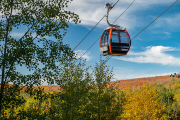 Cable car on a cable passes by nearby trees at a ski hill in Minnesota, with autumn colors on the distant hill and mostly blue sky.