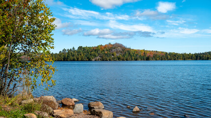 Beautiful blue water and sky at a lake in northern Minnesota on a sunny autumn day, with boulders along near shore and trees on rocky hill on the distant shoreline.