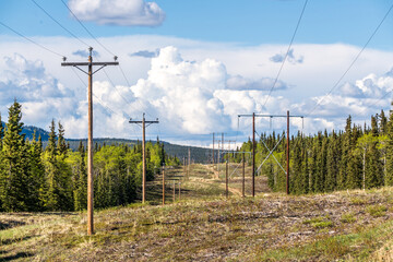 Electricity, power lines seen in rural Canada during summertime with blue sky, clouds and mountains, boreal forest landscape. 
