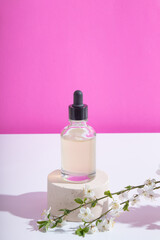 Obraz na płótnie Canvas Glass dropper bottle with serum or oil on podium with blooming twigs on pink background. Vertical stock photo with trendy hard shadows
