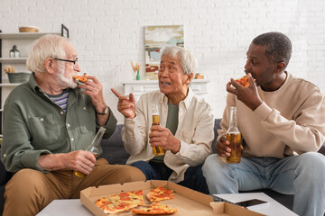 Senior asian man holding beer and pointing with finger near interracial friends eating pizza at home.