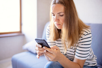 Young woman sitting, relaxing on sofa in living room, browsing wireless internet on smartphone, text message on modern mobile phone, shopping online through website