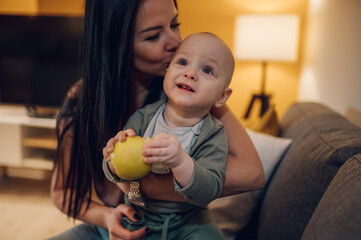 Fototapeta premium Baby boy sitting on the couch with his mom and eating an apple