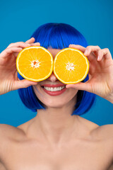 Healthy food and lifestyle concept. Beautiful woman with beige sun tan, smooth skin and blue wig holds cut orange in her hands near eyes and smiling with white teeth in vivid blue background