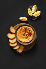 bowl of hummus with paprika on round wooden desk with pieces of bread and lemon on darck background, top view. copy space