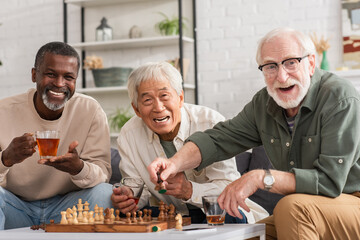 Cheerful multiethnic elderly friends looking at camera near chess and tea at home.