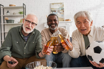 Positive interracial senior friends with football and beer looking at camera at home.